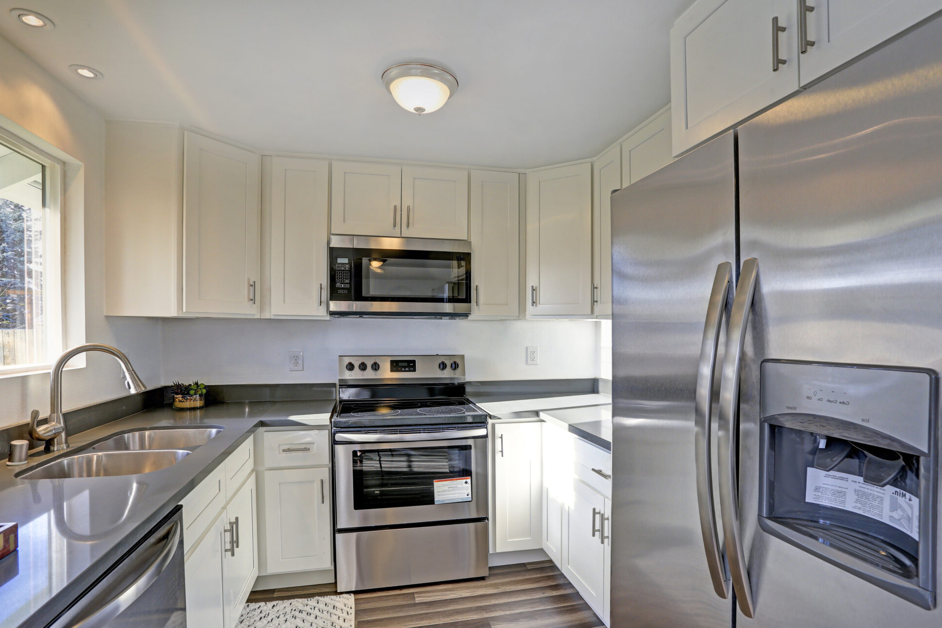 Light filled home interior features small compact kitchen with white cabinets and modern stainless steel appliances.