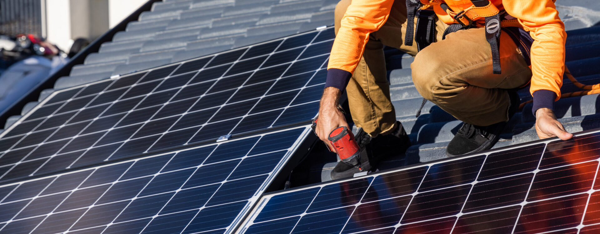 Solar panel technician with drill installing solar panels on house roof on a sunny day.