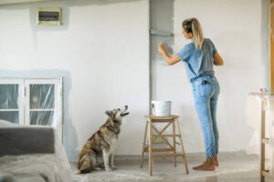 Woman painting with paint roller, she is dedicated and determined to finish her work right, dog is curious about what she doing