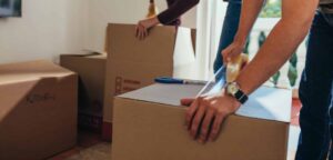 Close up of man sealing a packing box with adhesive tape while a woman placing articles in another box. Couple packing their items for moving into a new house in Nebraska.