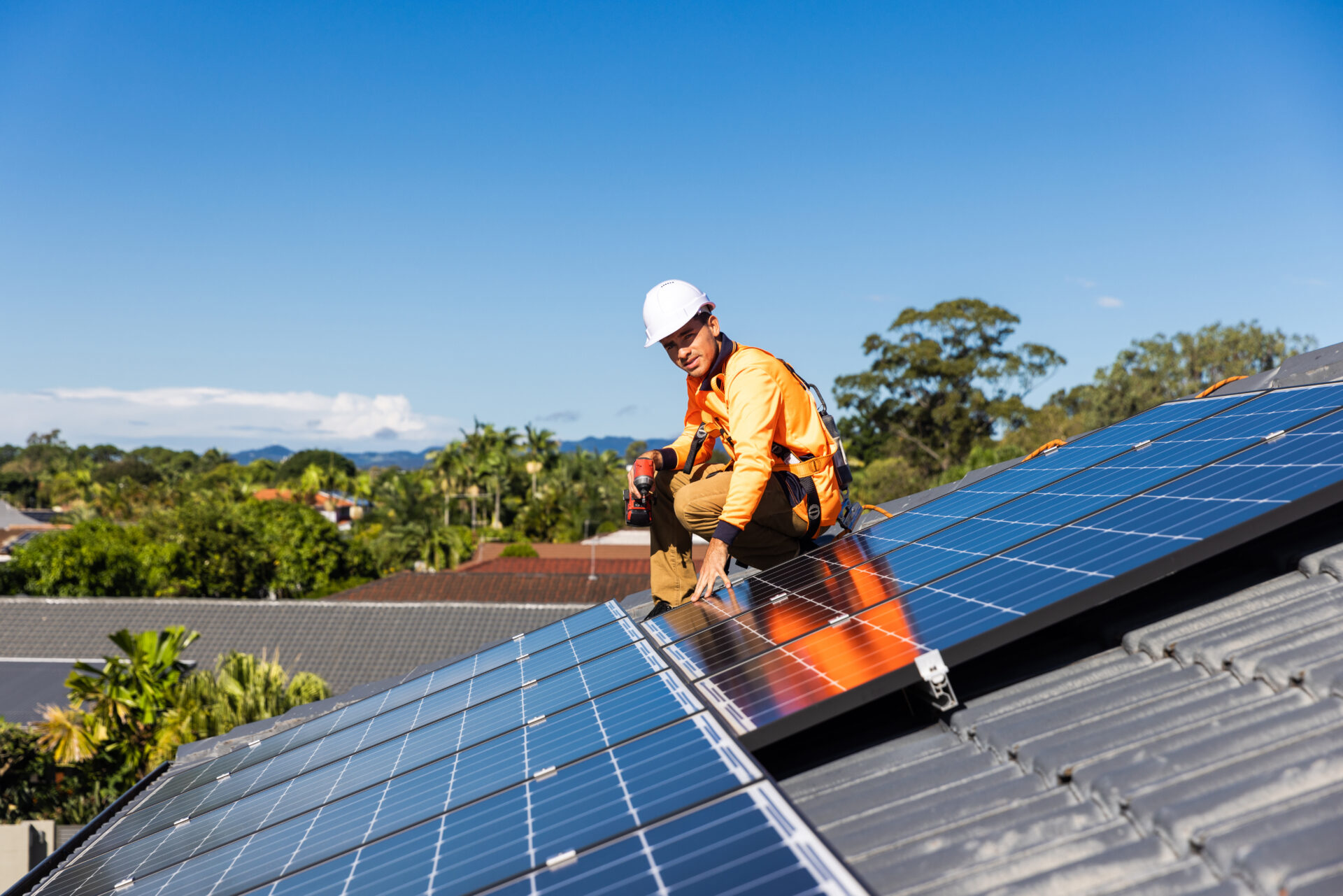 Solar panel technician with drill installing solar panels on house roof on a sunny day.