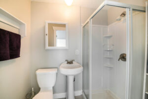 Interior of a small white bathroom with shower stall kit on the side. There is a mirror above the toilet beside the pedestal sink near the glass enclosure of the shower stall.