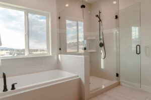 Built in bathtub with black faucet and shower stall with half glass enclosure. Interior of bathroom with view of snowy landscape and cloudy sky through the window.