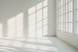 Sunlit empty room corner with large windows casting shadows on white walls and floor.