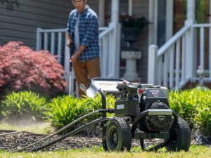 A person uses a pressure washer in a garden, surrounded by green shrubs and a red bush.