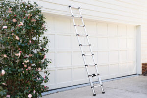 A silver telescopic ladder leans against a white garage door beside a blooming shrub with pink flowers.