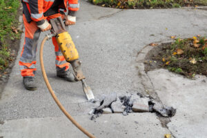 A worker in bright orange safety gear uses a yellow jackhammer on a cracked sidewalk, breaking concrete pieces.