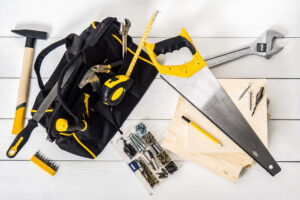 Toolkit spread on a light wooden surface, featuring a black bag, a yellow-handled saw, a hammer, wrench, tape measure, screws, and drill bits.
