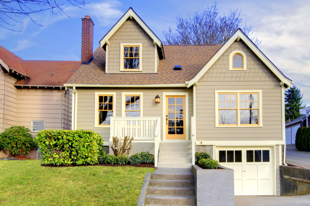 Charming beige house with dormer window and brick chimney. White trim, front steps, and garage. Manicured bushes and a sunny, inviting look.