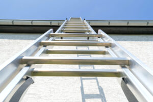 Tall silver ladder leaning against a white wall, reaching up to a building’s roof under a clear blue sky.