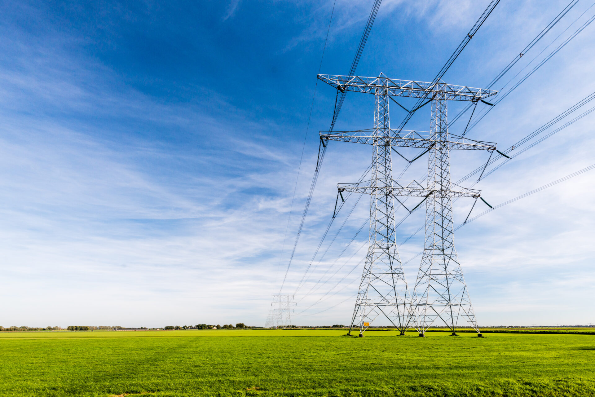 Hight voltage lines and power pylons in a flat and green agricultural landscape on a sunny day with cirrus clouds in the blue sky.