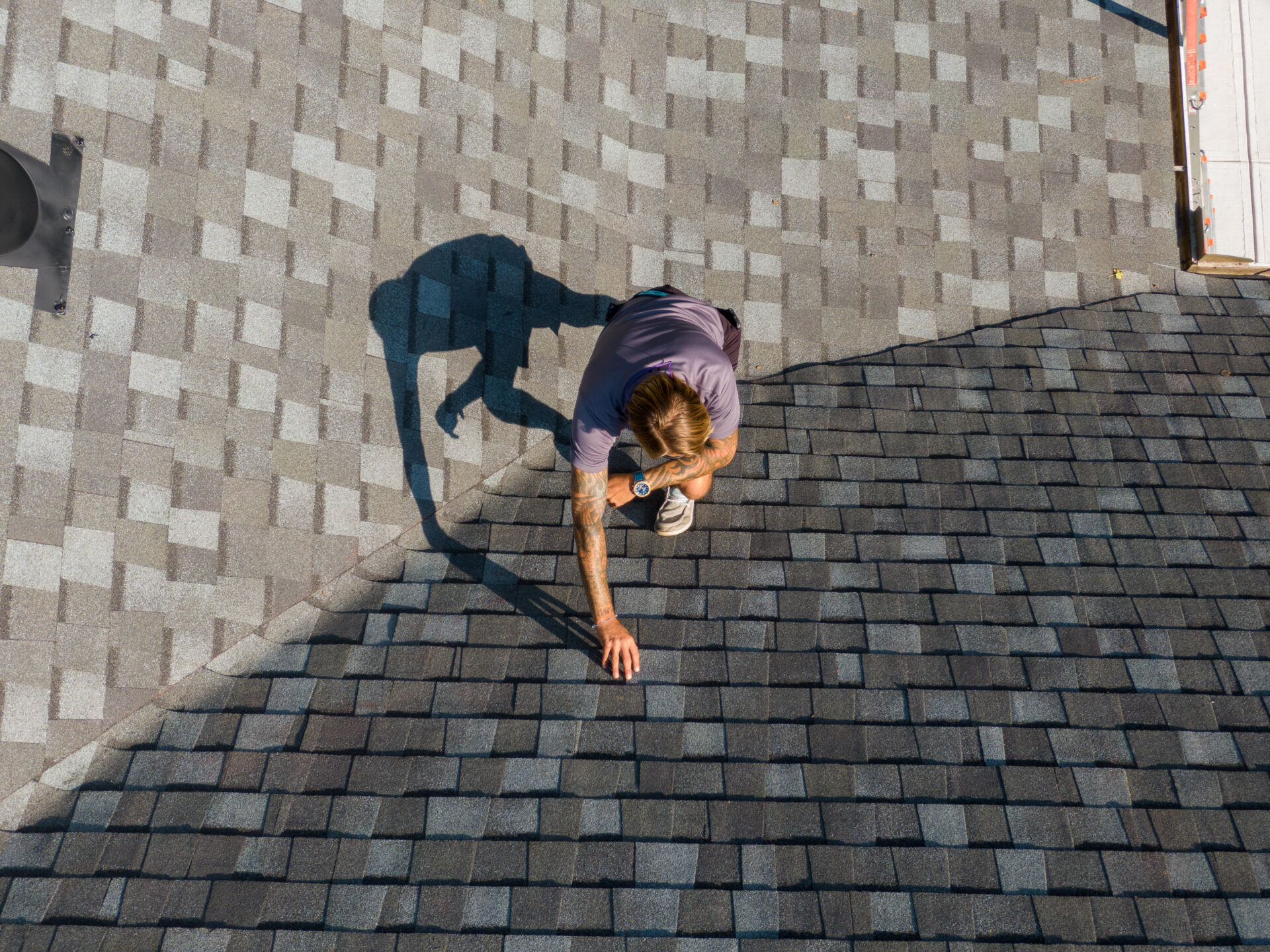 A person in casual clothing carefully inspecting a rooftop with grey shingles, casting a distinct shadow.