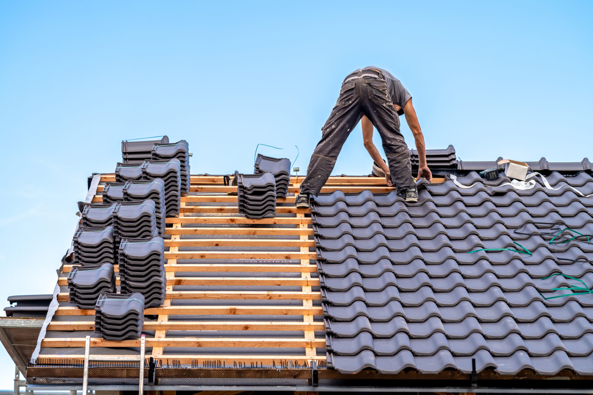 A worker installs black clay roof tiles on a house under a clear blue sky.