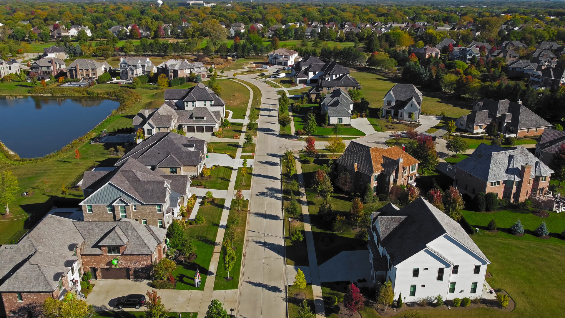 Aerial view of suburban neighborhood in Chicago, Illinois. A scenic aerial view of a suburban neighborhood in Chicago, Illinois, featuring large homes with manicured lawns, a curved street, and a tranquil pond surrounded by trees in autumn colors.