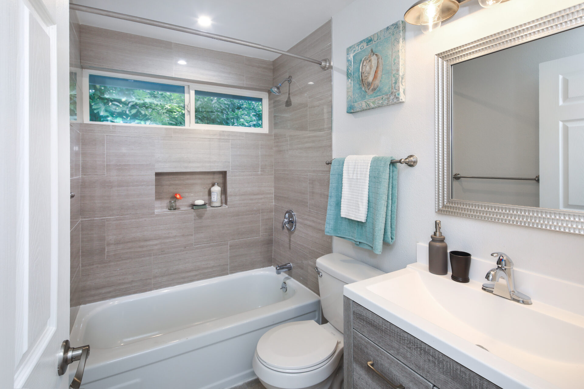 Modern bathroom with gray tiles, bathtub, and mounted shower. A sink with a large mirror, teal towels, and a seashell artwork add a fresh feel.