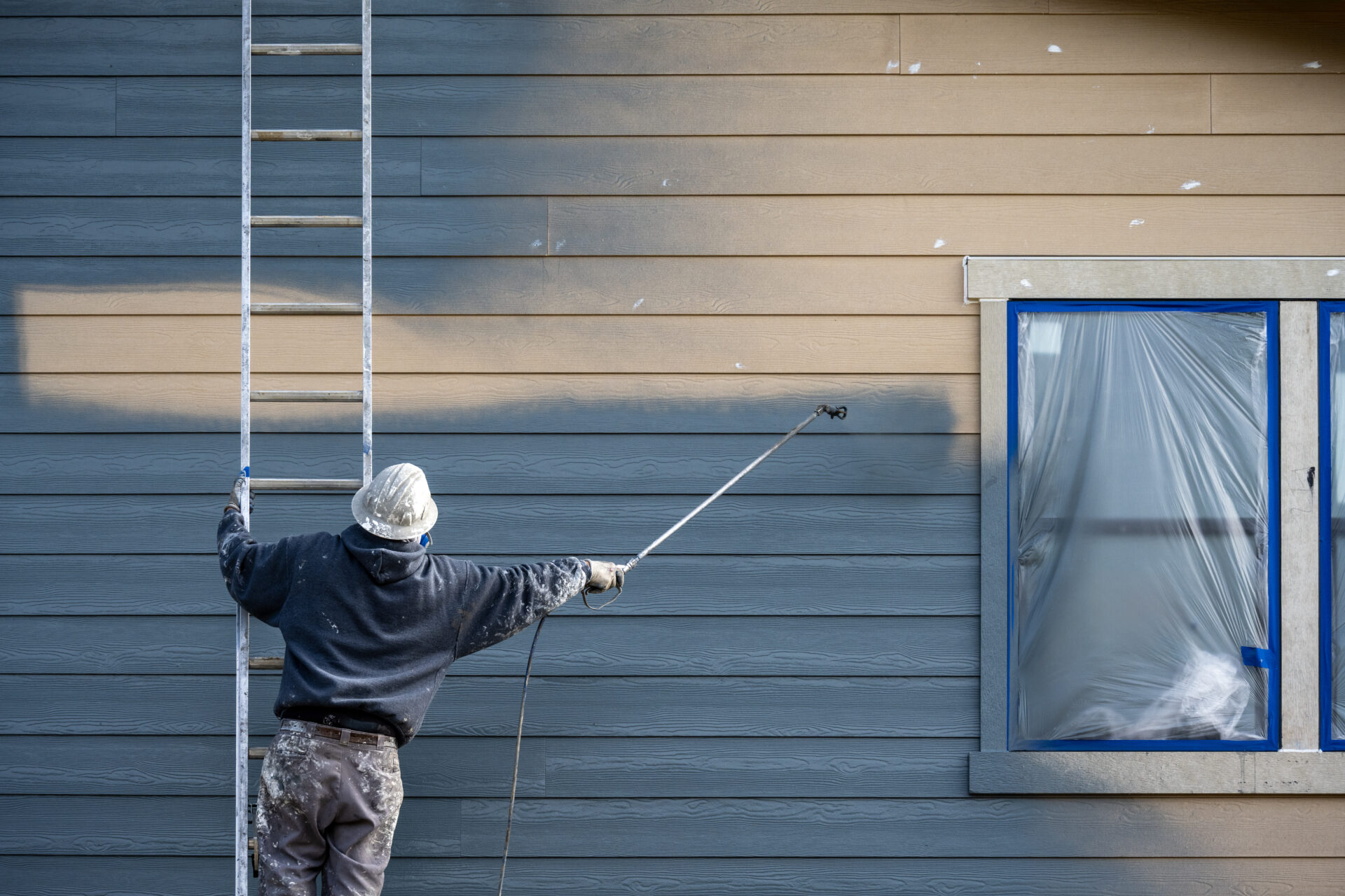 Professional painter in hard hat and air respirator on extension ladder with paint sprayer painting back exterior of house.